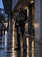 Homme portant l'ensemble jogging noir à strass Hype Or Nothing DIFFERENT, sweat à capuche et pantalon à bandes, sous une galerie la nuit – DIFFERENT Paris