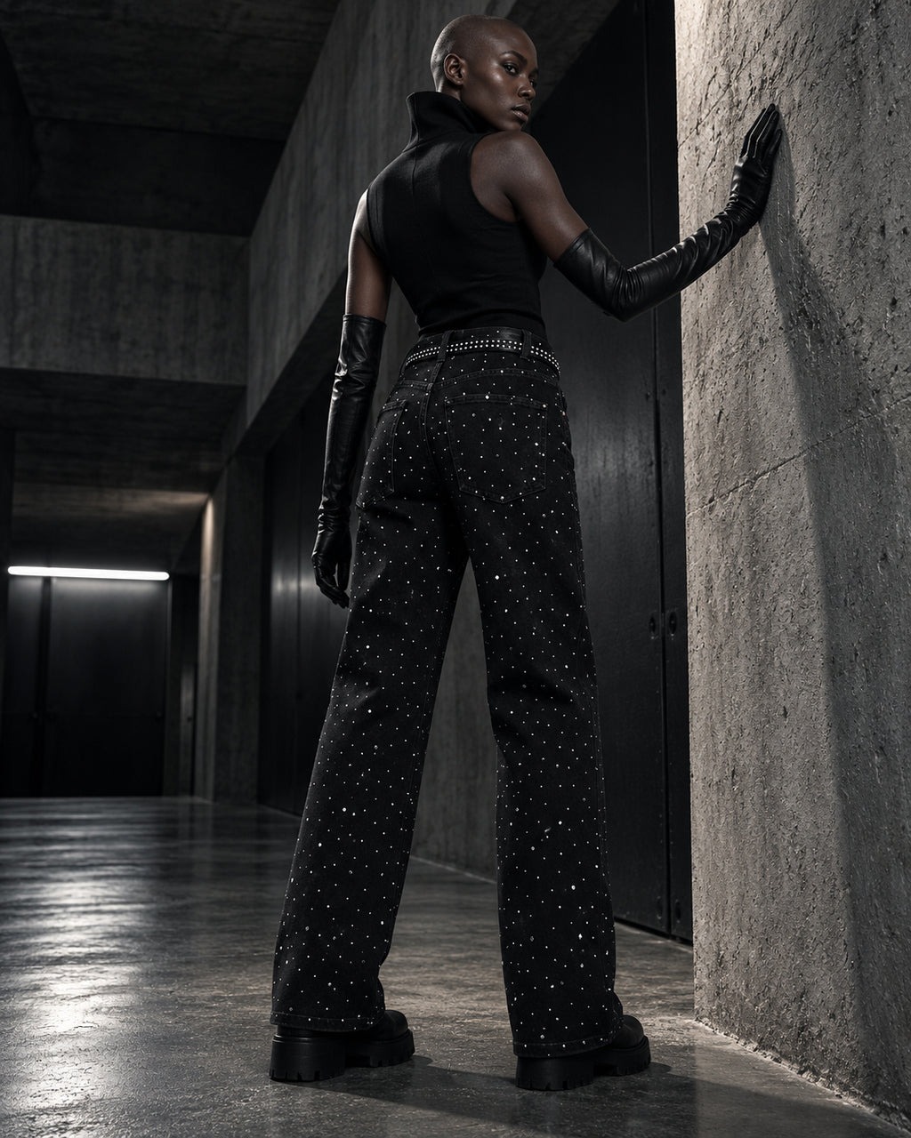 Femme de dos portant le jean à strass noir DIFFERENT avec gants longs, appuyée contre un mur en béton – DIFFERENT Paris