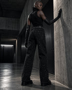 Femme de dos portant le jean à strass noir DIFFERENT avec gants longs, appuyée contre un mur en béton – DIFFERENT Paris
