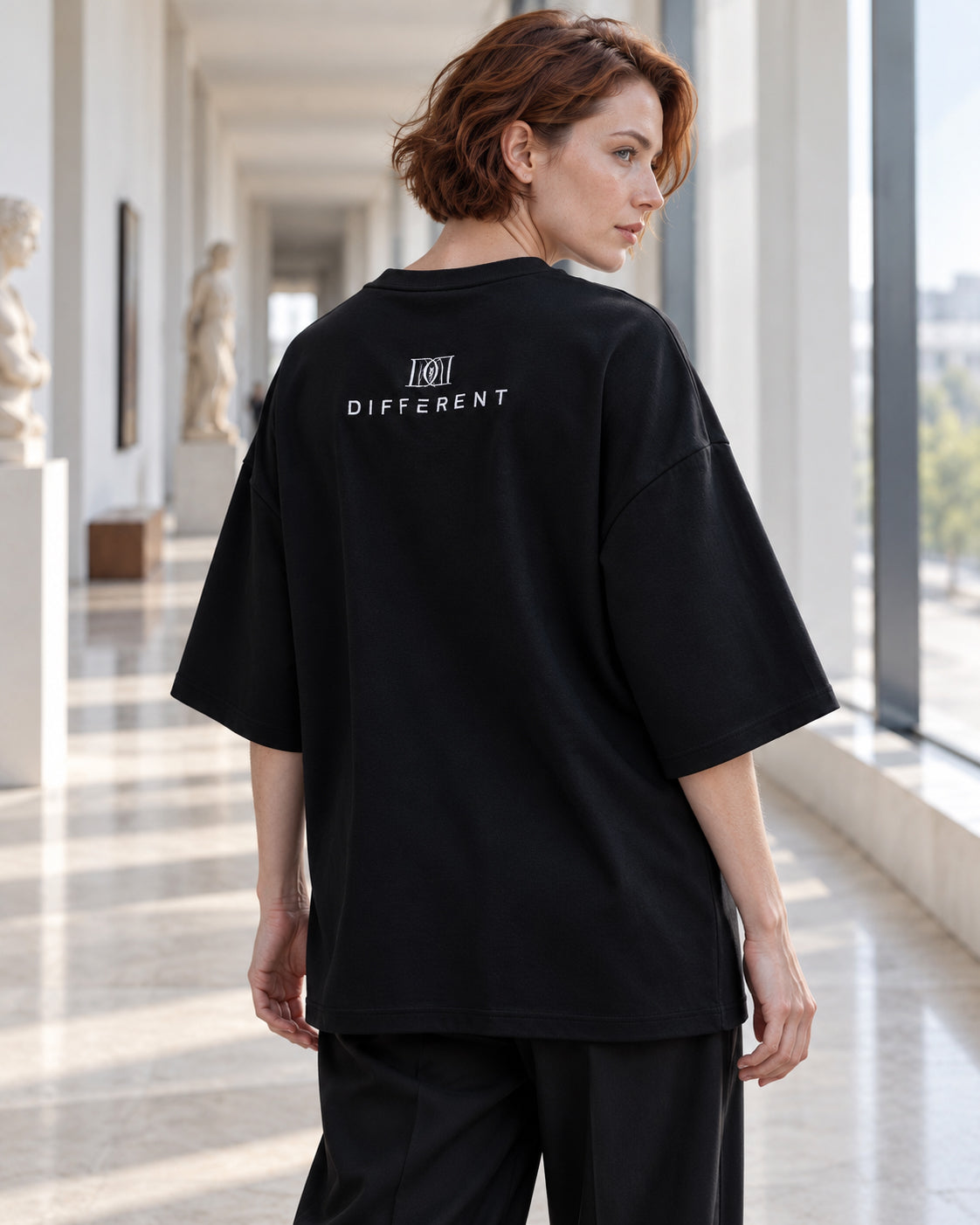 Femme de dos portant le t-shirt oversize premium noir avec logo DIFFERENT dans le dos, dans une galerie lumineuse – DIFFERENT Paris