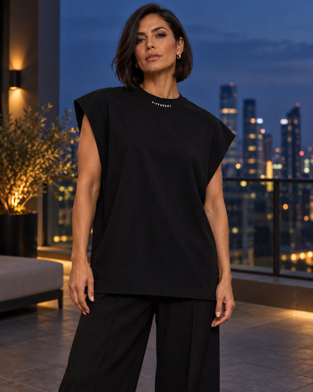 Femme portant le t-shirt sans manches oversize noir DIFFERENT avec pantalon large noir, sur une terrasse de nuit avec vue sur la ville – DIFFERENT Paris