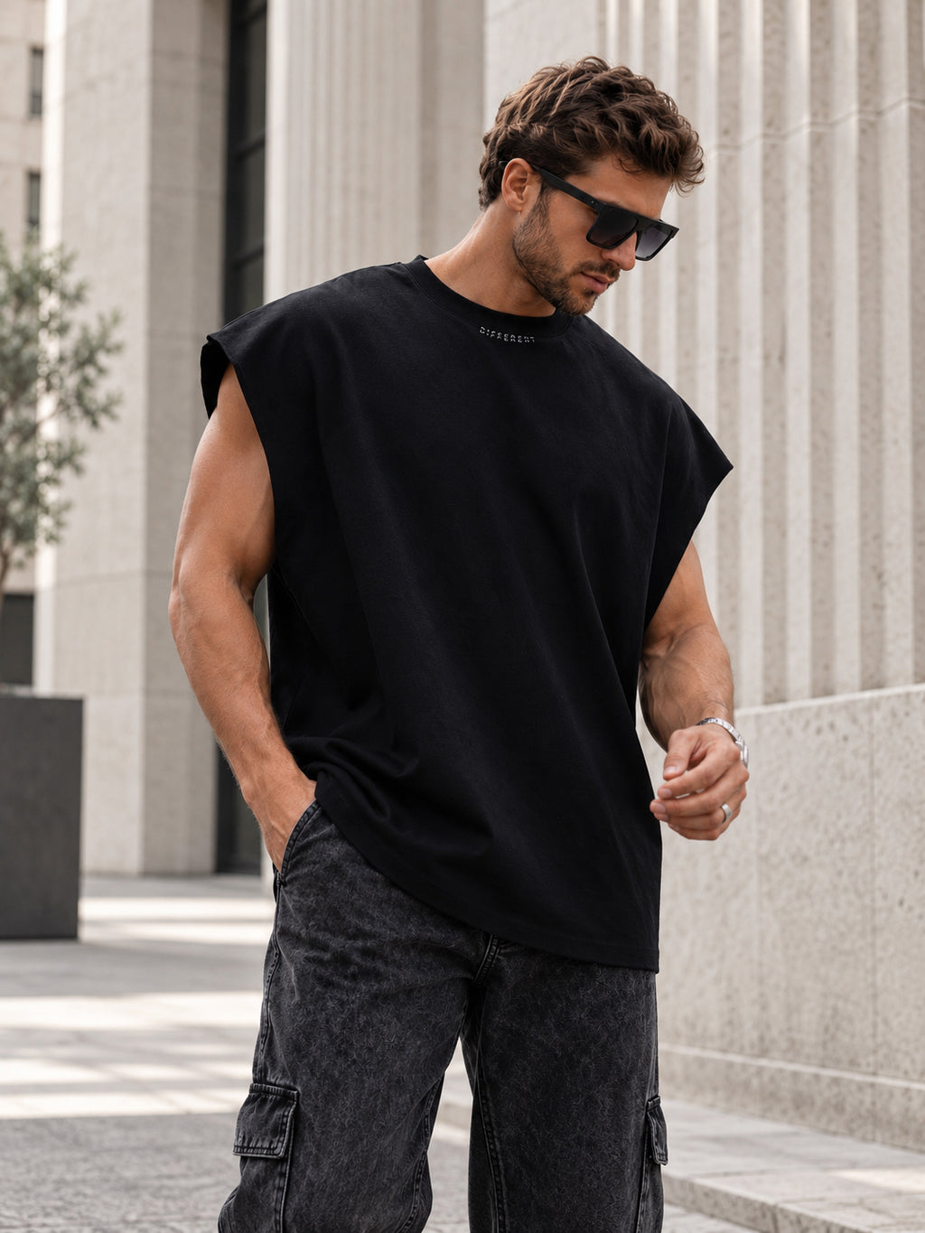 Homme portant le t-shirt sans manches oversize noir DIFFERENT avec jean cargo gris et lunettes de soleil, devant des colonnes – DIFFERENT Paris