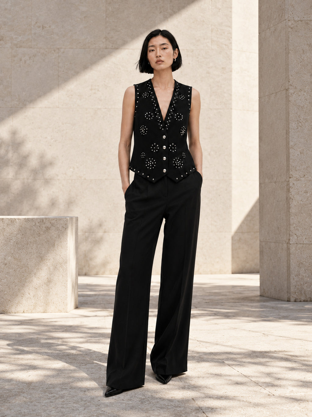 Femme portant le gilet noir structuré à clous argentés et diamants DIFFERENT avec pantalon large noir, dans une cour ensoleillée – DIFFERENT Paris