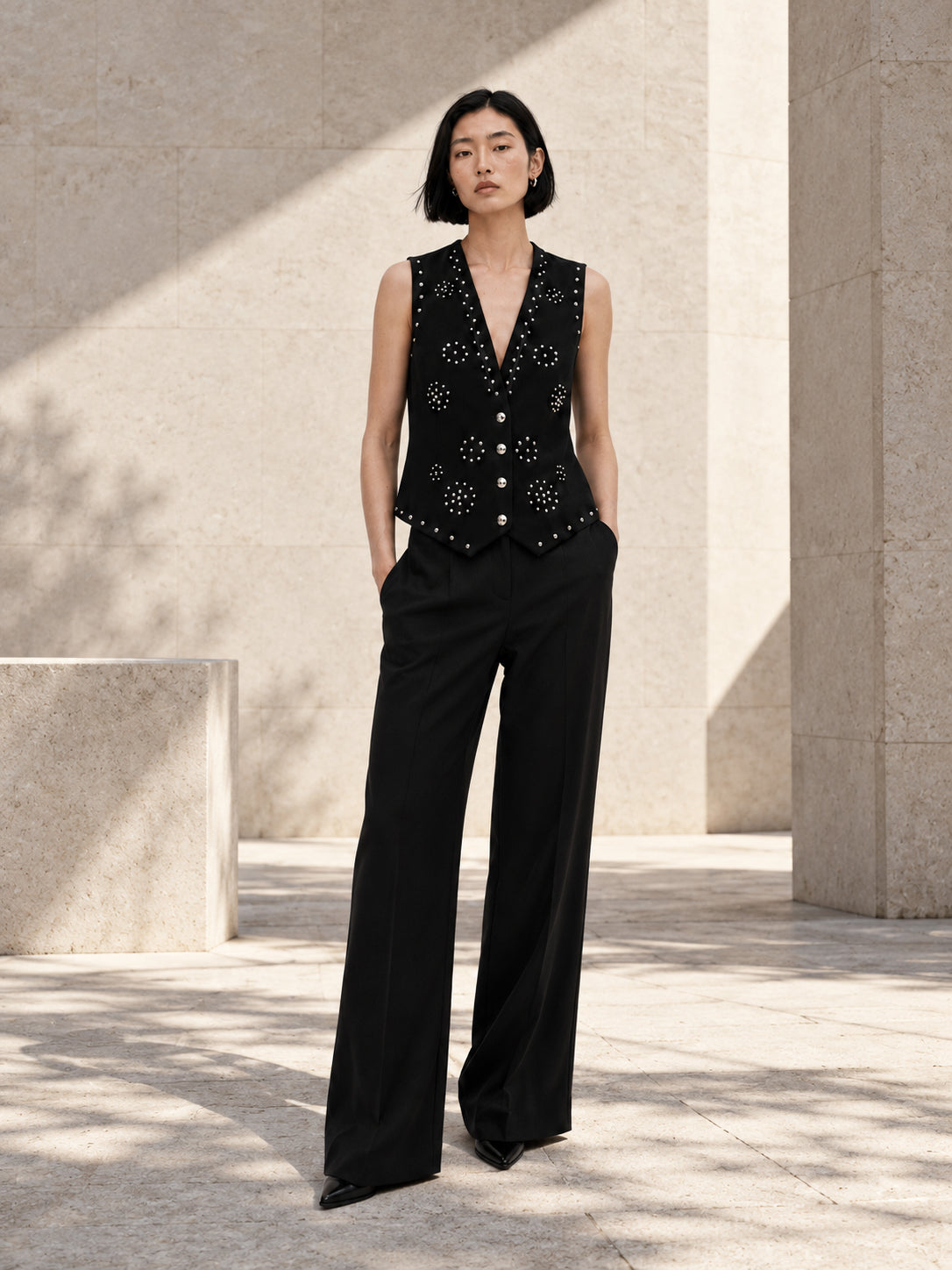 Femme portant le gilet noir structuré à clous argentés et diamants DIFFERENT avec pantalon large noir, dans une cour ensoleillée – DIFFERENT Paris