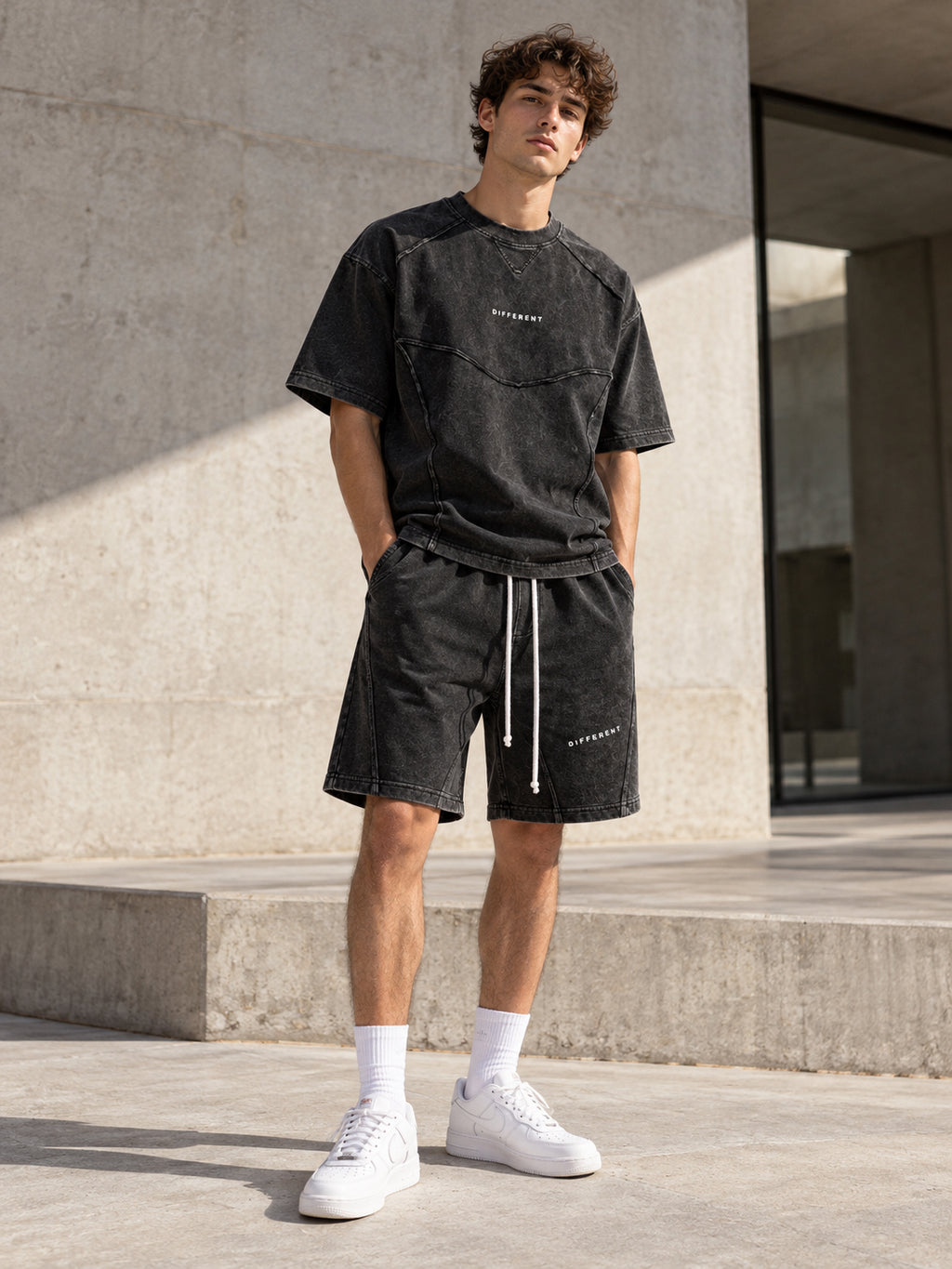 Homme portant l'ensemble t-shirt et short oversize gris délavé DIFFERENT avec baskets blanches, devant un mur en béton – DIFFERENT Paris