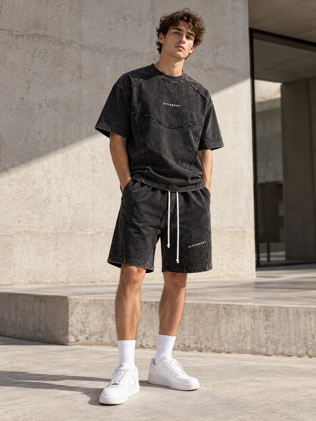 Homme portant l'ensemble t-shirt et short oversize gris délavé DIFFERENT avec baskets blanches, devant un mur en béton – DIFFERENT Paris