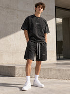 Homme portant l'ensemble t-shirt et short oversize gris délavé DIFFERENT avec baskets blanches, devant un mur en béton – DIFFERENT Paris
