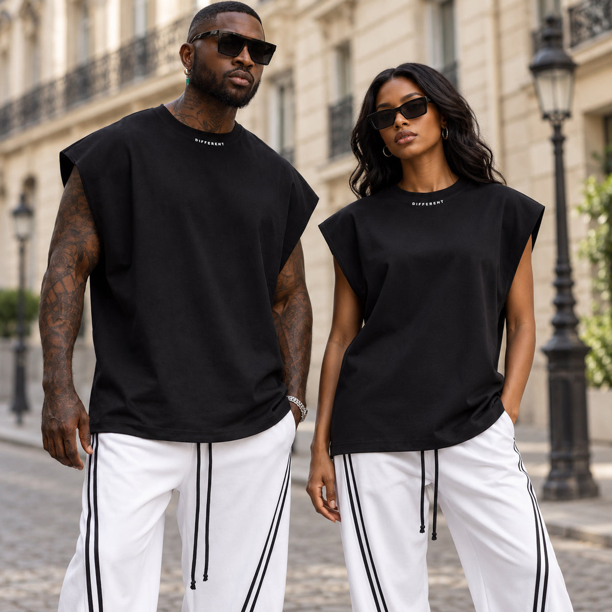 Homme et femme portant le t-shirt sans manches oversize noir DIFFERENT en coton épais 260 g avec pantalon blanc à bandes, dans une rue de Paris – DIFFERENT Paris
