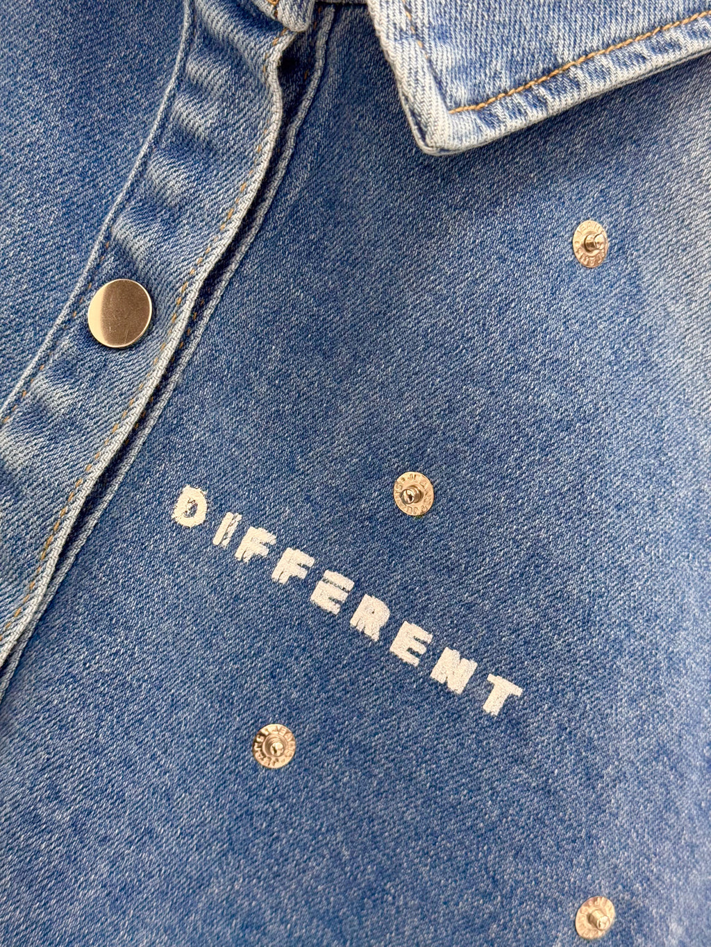 Gros plan sur la veste denim bleue DIFFERENT avec logo blanc imprimé, boutons pression métalliques et rivets décoratifs