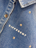 Gros plan sur la veste denim bleue DIFFERENT avec logo blanc imprimé, boutons pression métalliques et rivets décoratifs