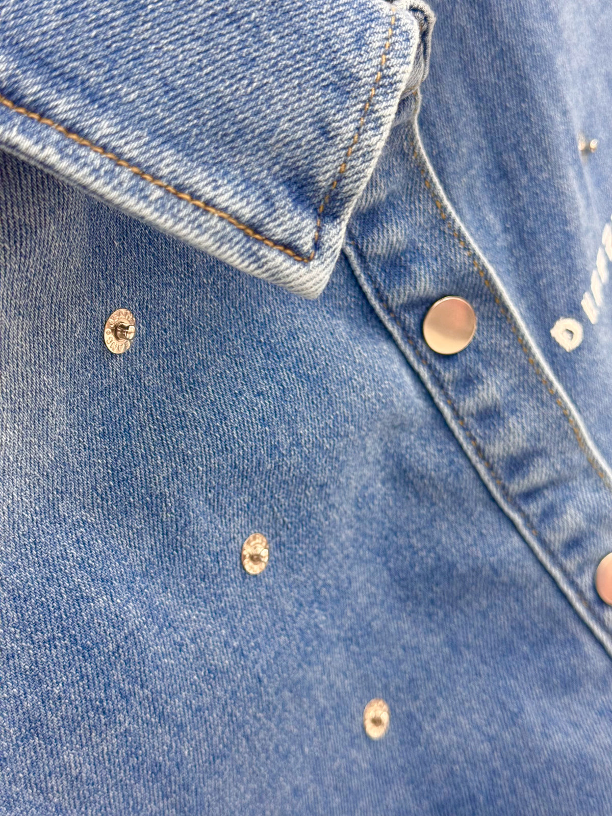 Détail de la veste courte en denim bleu DIFFERENT avec col chemise, boutons métalliques et rivets décoratifs.