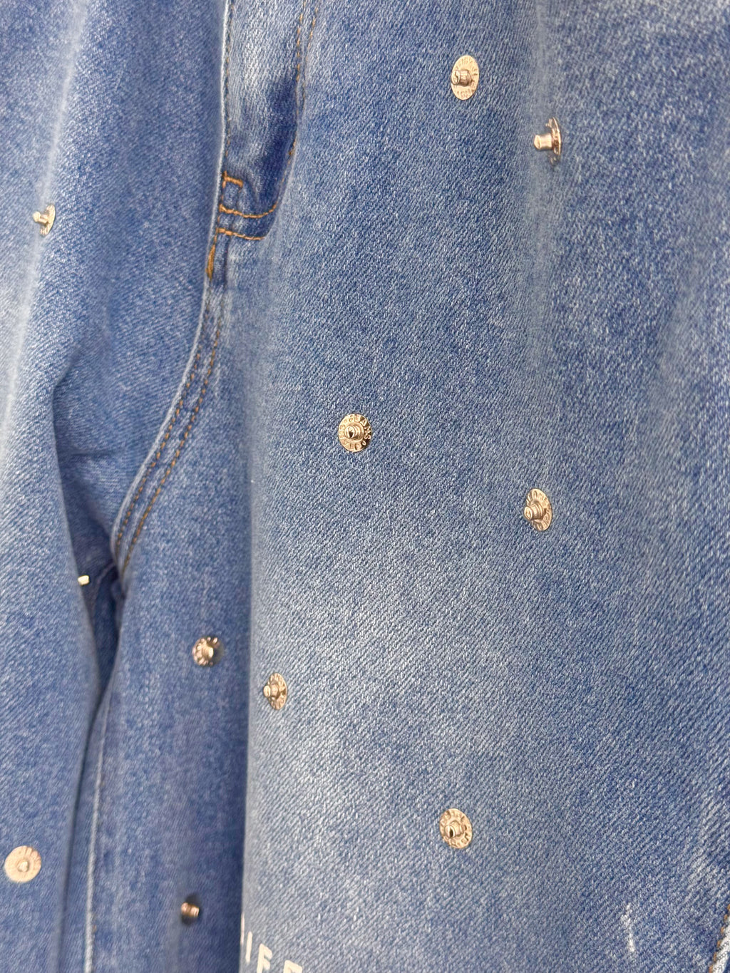 Gros plan sur denim bleu DIFFERENT avec texture délavée, surpiqûres contrastées et petits rivets métalliques décoratifs.