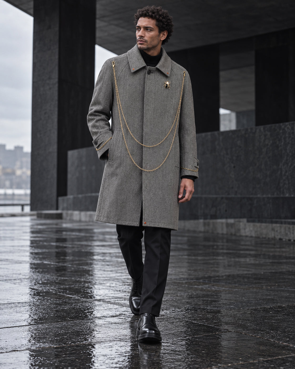 Homme portant le manteau long gris à chaînes dorées DIFFERENT, marchant sur une esplanade mouillée – DIFFERENT Paris