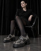 Femme assise en tenue noire et collants noirs portant les chaussures à plateforme motif baroque DIFFERENT – DIFFERENT Paris