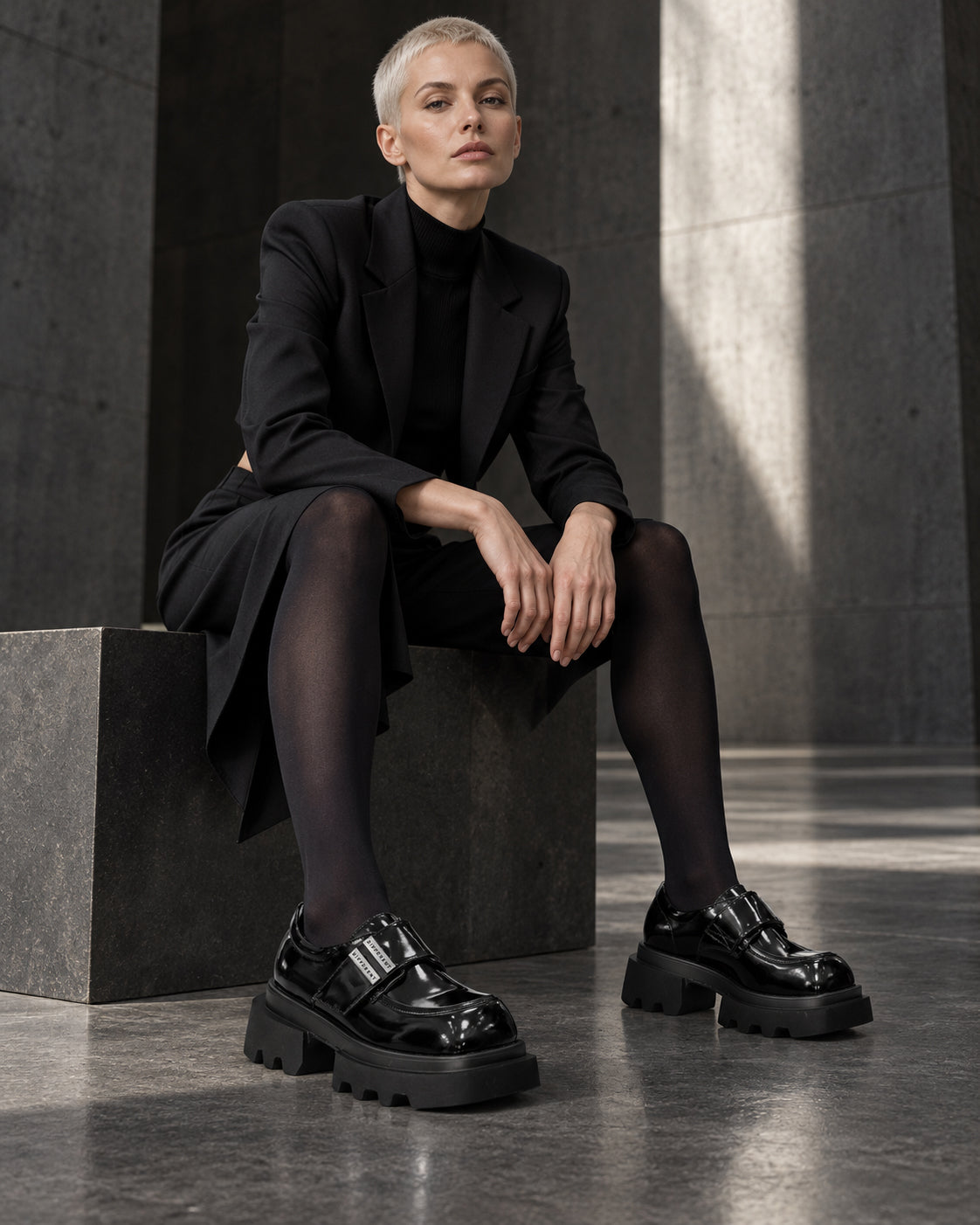 Femme assise en manteau noir et collants noirs portant les chaussures plateforme noir brillant DIFFERENT – DIFFERENT Paris