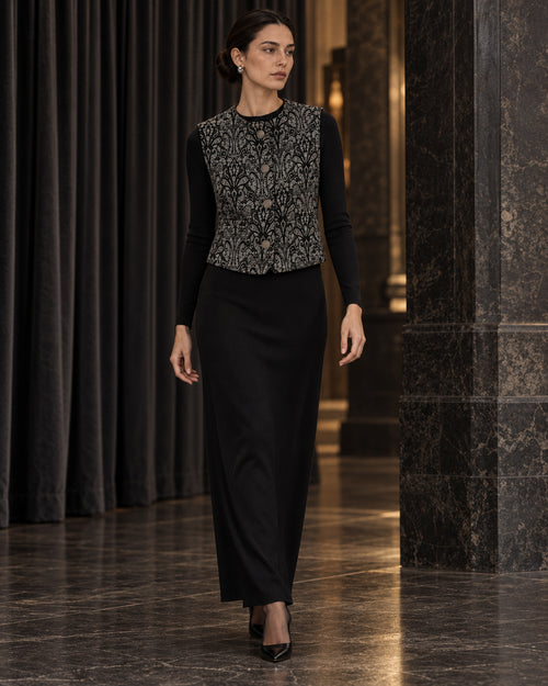 Femme portant le gilet court à motif baroque noir et gris DIFFERENT avec haut noir et jupe longue noire, dans un couloir – DIFFERENT Paris