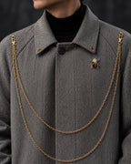 Gros plan sur le col du manteau long gris avec chaînes dorées en double rang et broche bijou – DIFFERENT Paris