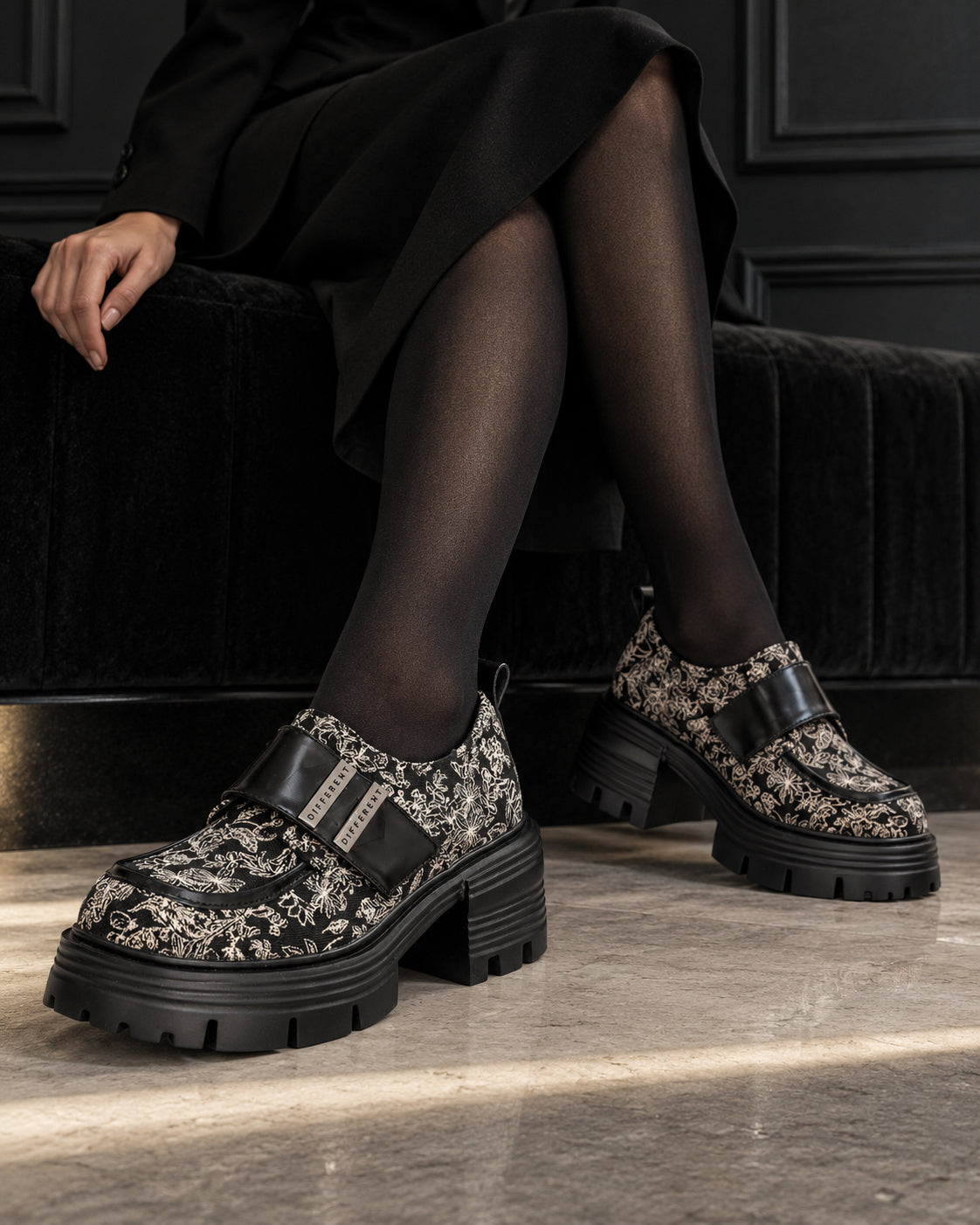 Chaussures à plateforme motif baroque DIFFERENT portées avec robe noire et collants noirs, jambes croisées – DIFFERENT Paris