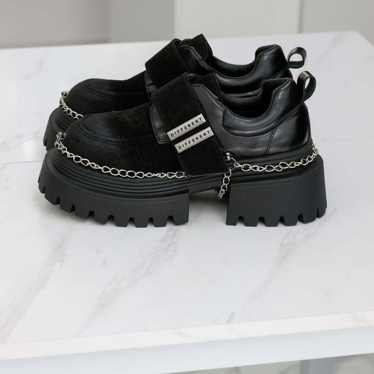 Paire de chaussures noires vue de côté, avec talon large, chaînes argentées et languettes à l’arrière.