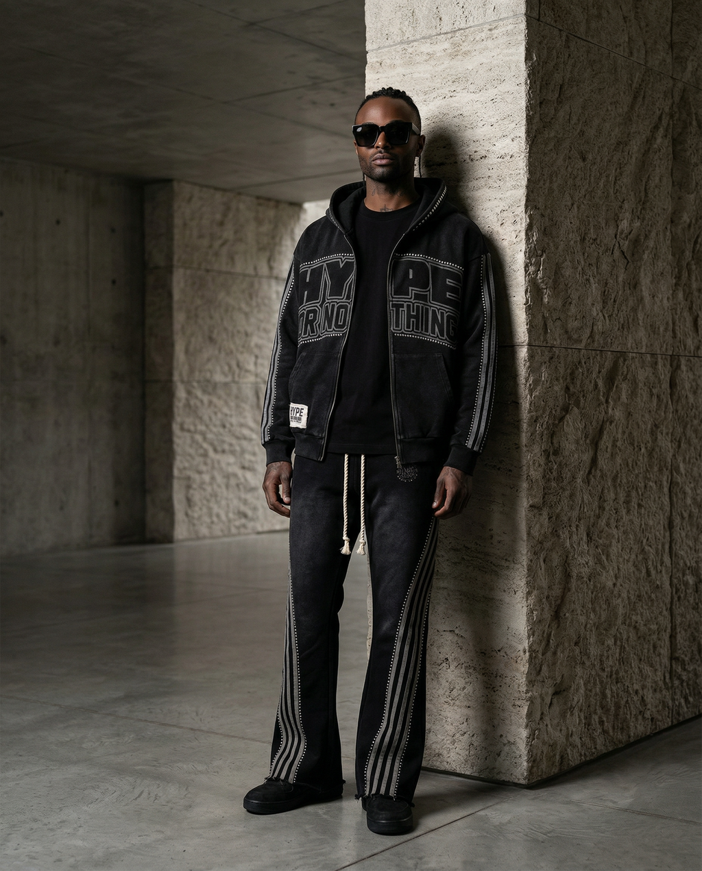 Black HN jogging set worn by a man, open Hype Or Nothing zip-up hoodie and rhinestone striped trousers, concrete backdrop – DIFFERENT Paris