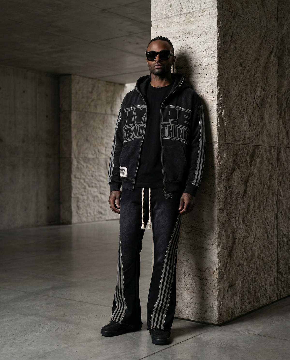 Black HN jogging set worn by a man, open Hype Or Nothing zip-up hoodie and rhinestone striped trousers, concrete backdrop – DIFFERENT Paris